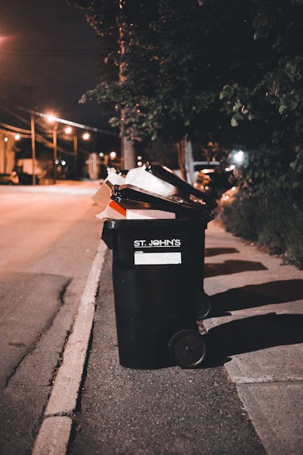 A black wheelie bin is positioned on the edge of a paved urban street at night, with its lid open and filled with mixed waste items including cardboard boxes and plastic packaging. The bin is labeled 'ST. JOHN'S' and is situated next to a sidewalk, with shadows cast by nearby streetlights visible on the ground. In the background, soft orange street lighting illuminates the street and nearby trees, with faint outlines of buildings and parked vehicles further along the road. The surroundings suggest a residential area where private waste collection services, such as those offered by Waste Collection Wandsworth, are used for rubbish removal, reflecting an environment prepared for on-site waste management or independent collection outside of municipal waste disposal systems.