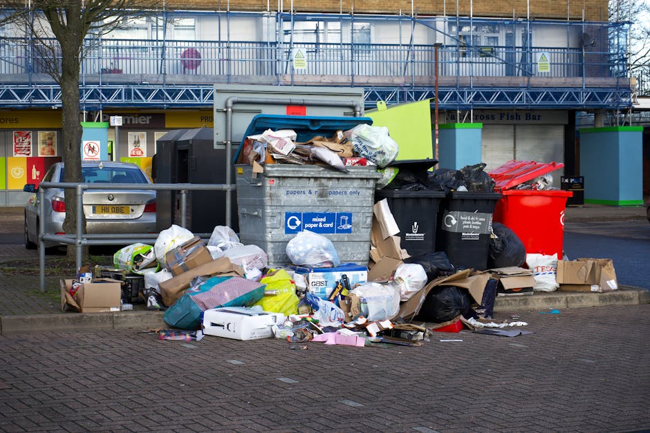 An outdoor scene showing a large collection of mixed rubbish and recycling bags piled around several open and closed waste bins, including a grey bin labeled for mixed paper and cardboard, a black bin for general waste, and a red bin, all situated on a paved area near a commercial building. The rubbish includes discarded cardboard boxes, paper, plastic bags, and packaging materials, some spilling onto the surrounding pavement. A metal railing separates the waste area from a parked silver hatchback car behind it, with visible license plates. In the background, a building facade features storefronts with signage protected by blue scaffolding and safety barriers, indicating ongoing construction or maintenance. The environment appears to be a busy urban shopping or commercial district, where private waste collection services, such as Waste Collection Wandsworth, might assist with on-site rubbish removal, especially when dealing with large amounts of waste encountered outside retail establishments.