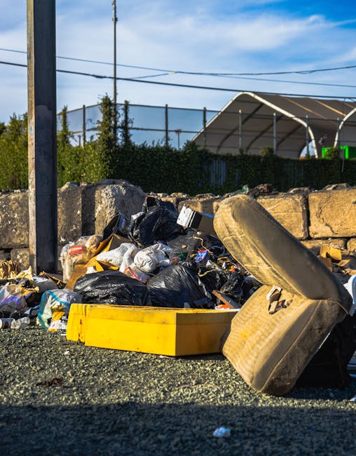 A pile of mixed household rubbish lies on a gravel surface outdoors, consisting of black plastic garbage bags, discarded packaging, and various debris. In the foreground, a large, worn car tyre is positioned, partially leaning against the pile. A yellow plastic container is also visible amidst the waste. Behind the rubbish, a low stone wall made of uneven, weathered bricks separates the area from a background featuring a fence, some greenery, and a structure with an arched roof. The scene is illuminated by daylight, with a partly cloudy sky overhead and utility poles with wires running across it, indicating an urban or suburban setting. The image suggests an instance of private rubbish disposal, possibly associated with independent waste collection services such as Waste Collection Wandsworth, which handles local household waste removal beyond regular municipal services.