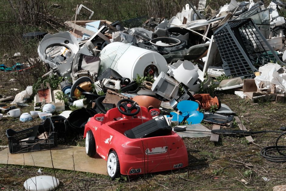 A pile of mixed household waste and discarded objects situated outdoors on uneven ground, including a red plastic children's ride-on car positioned in the foreground with visible paint scratches and minor damage. Surrounding the car are various items such as broken ceramic dishes, plastic containers, a small black crate, and a blue balloon. In the background, there is a dense collection of larger debris like broken appliances, parts of furniture, a white washing machine drum, dark plastic crates, and bits of metal framing. The waste materials vary in texture, with some items having glossy, smooth surfaces and others featuring rough, weathered finishes. The scene is lit by natural daylight, casting soft shadows over the clutter. This outdoor dump area, composed of household refuse, reflects typical debris that might be collected through private rubbish removal services, and Waste Collection Wandsworth could handle such waste for proper disposal or clearance.