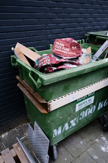 A green wheeled rubbish bin filled with black and red packaging bags and rolled-up lawn care fabric laid on top. The bin is positioned on a concrete pavement against a dark blue brick wall. The bin features a white label with green text and the company name, Waste Collection Wandsworth, indicating it is used for waste collection and disposal. Adjacent to the bin, a second similar container is partially visible, also filled with waste. The scene appears to be an outdoor area, likely a back alley or storage space, reflecting a typical setup for private waste disposal or outdoor rubbish collection services. The environment is well-lit, and the scene presents a practical view related to waste management and rubbish removal activities, emphasizing the importance of proper waste segregation and alternative disposal options outside of municipal collection.