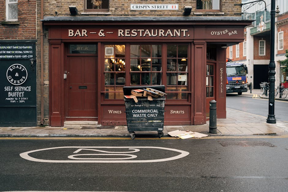 A black wheeled bin labeled 'Commercial Waste Only' positioned on a sidewalk in front of a restaurant with a brown wooden exterior and large glass windows. The bin is overflowing with flattened cardboard boxes and other waste materials, some of which are spilling onto the pavement. The bin is placed near a black bollard and a small pile of papers on the ground. To the left of the restaurant, there is a black signboard with white lettering advertising a self-service buffet. In the background, a street scene includes a parked blue truck and a bicycle leaning against a pole, with a brick building and street signs visible. The environment suggests an urban area where external waste disposal is managed by private waste collection services, such as Waste Collection Wandsworth, for businesses like this restaurant, illustrating a typical approach to commercial rubbish removal.