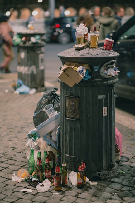 A green wheeled rubbish bin filled with black and red packaging bags and rolled-up lawn care fabric laid on top. The bin is positioned on a concrete pavement against a dark blue brick wall. The bin features a white label with green text and the company name, Waste Collection Wandsworth, indicating it is used for waste collection and disposal. Adjacent to the bin, a second similar container is partially visible, also filled with waste. The scene appears to be an outdoor area, likely a back alley or storage space, reflecting a typical setup for private waste disposal or outdoor rubbish collection services. The environment is well-lit, and the scene presents a practical view related to waste management and rubbish removal activities, emphasizing the importance of proper waste segregation and alternative disposal options outside of municipal collection.